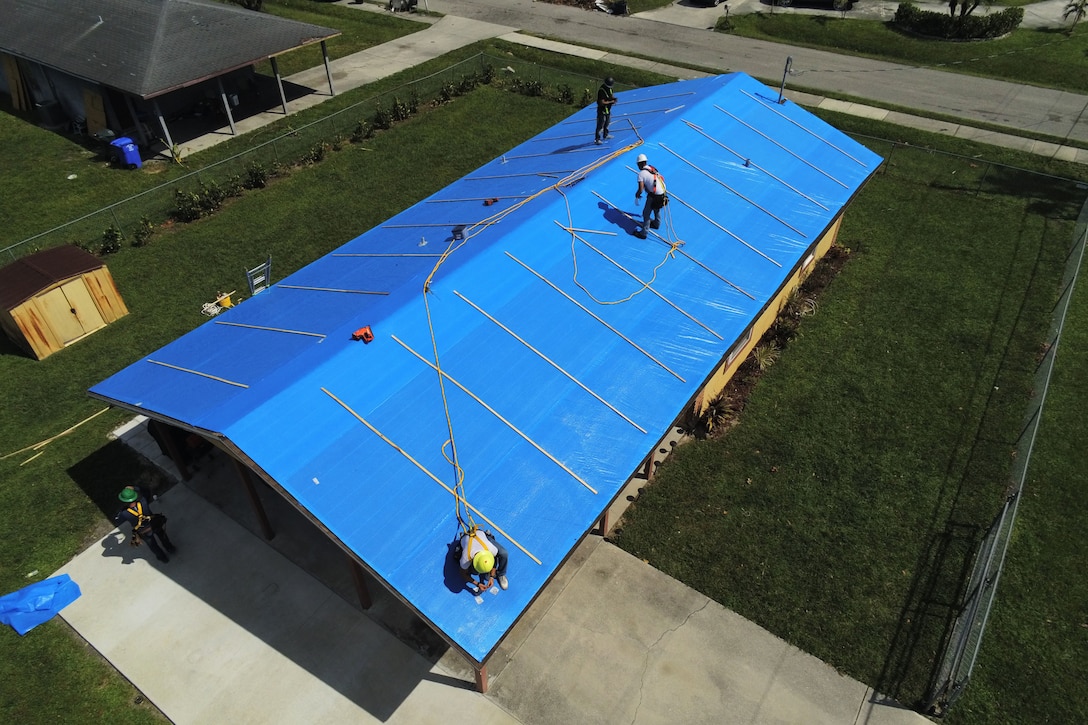 Three men lay a blue protective sheet on the roof of a home.