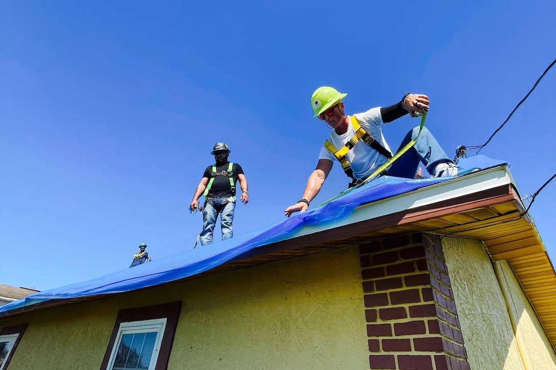 Three men lay a blue protective sheet on the roof of a home.