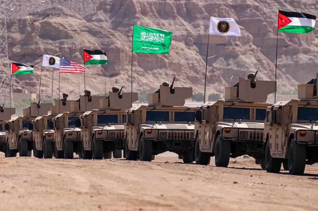 Humvees displaying flags drive in a close line along desert-like terrain.