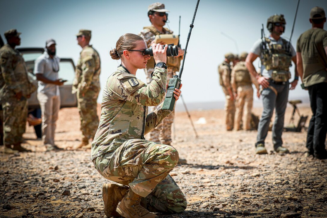 A soldier looks through binoculars in a desert-like area as service members work nearby.