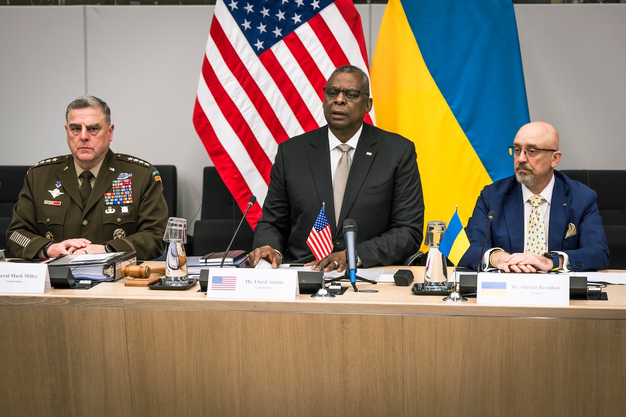 Three men, one in a military uniform, are seated at a table with microphones. Flags of the United States and Ukraine are in the background.