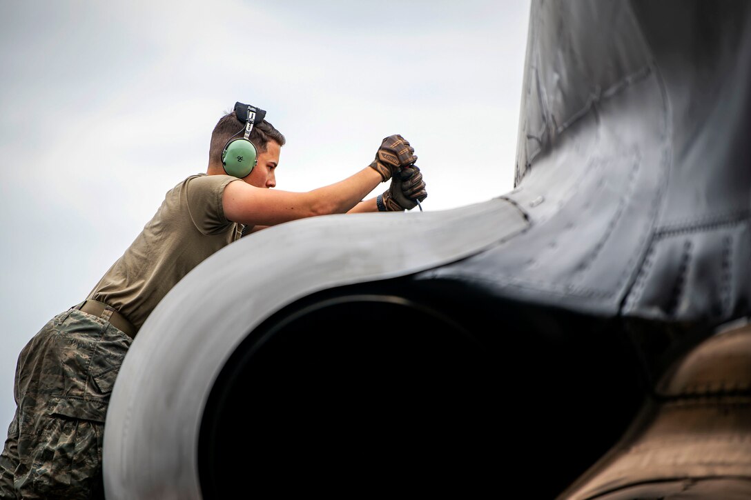 An airman wears headphones and work gloves while using a tool on the engine of an aircraft.