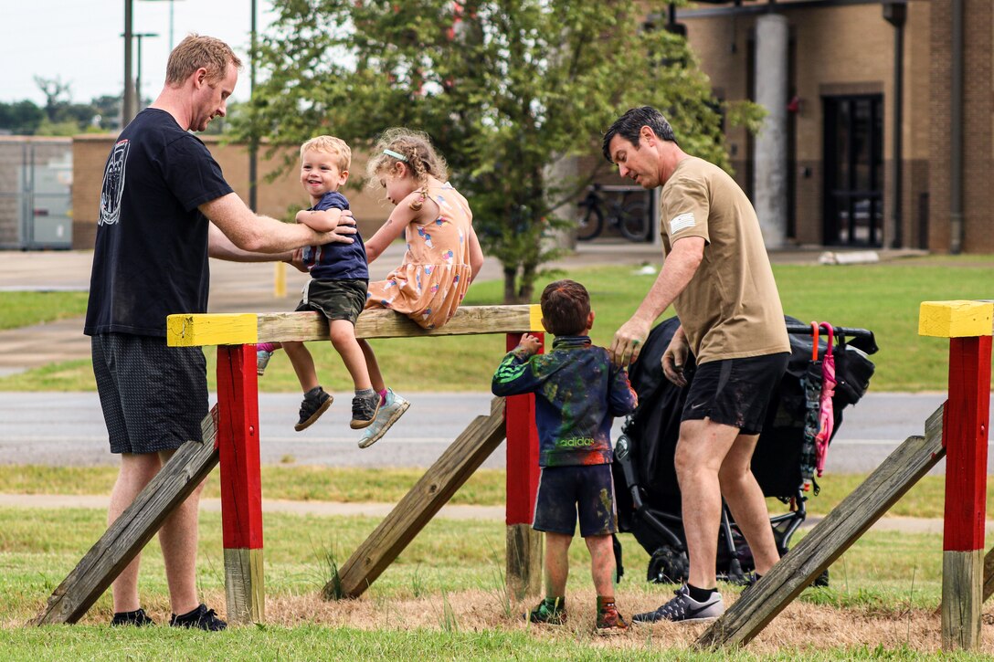 An adult holds a young child on a wooden obstacle where another kid sits; another adult and child stand nearby.