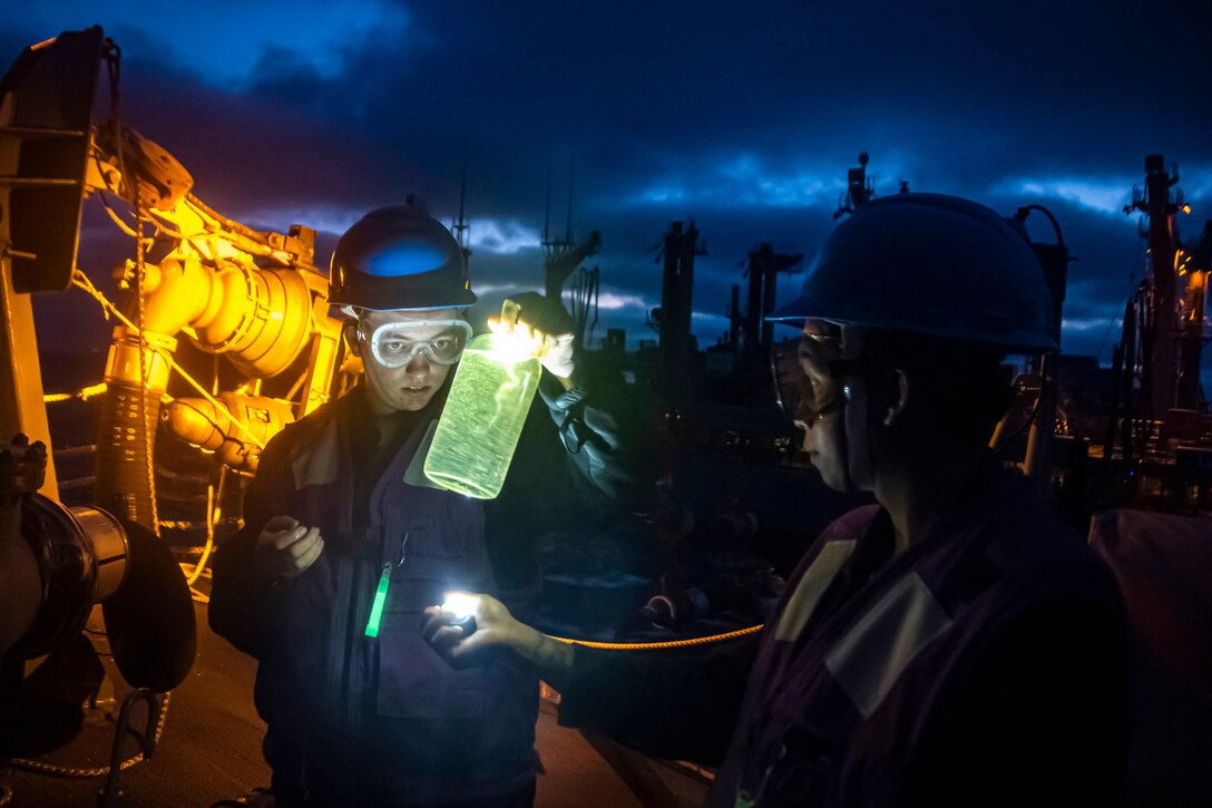 A sailor holds a light to a bottle a fellow sailor is holding aboard a ship at sea.