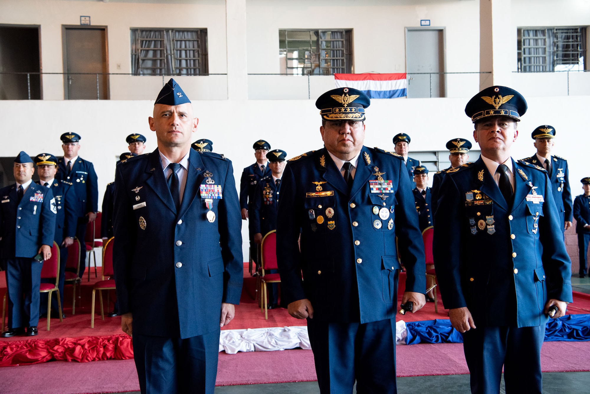 Three men stand side-by-side, all standing at attention. A group of military members are standing behind them.