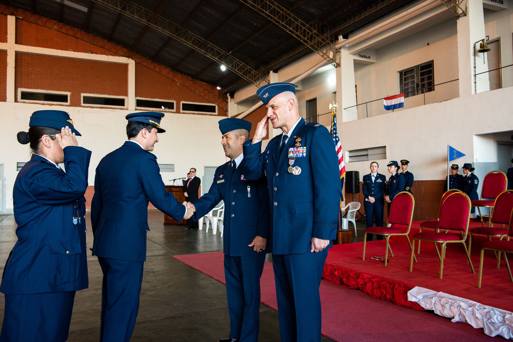 Four people frame the photo; the woman on the left is saluting the man positioned at the right side. The two people in the middle, both men, are shaking hands.