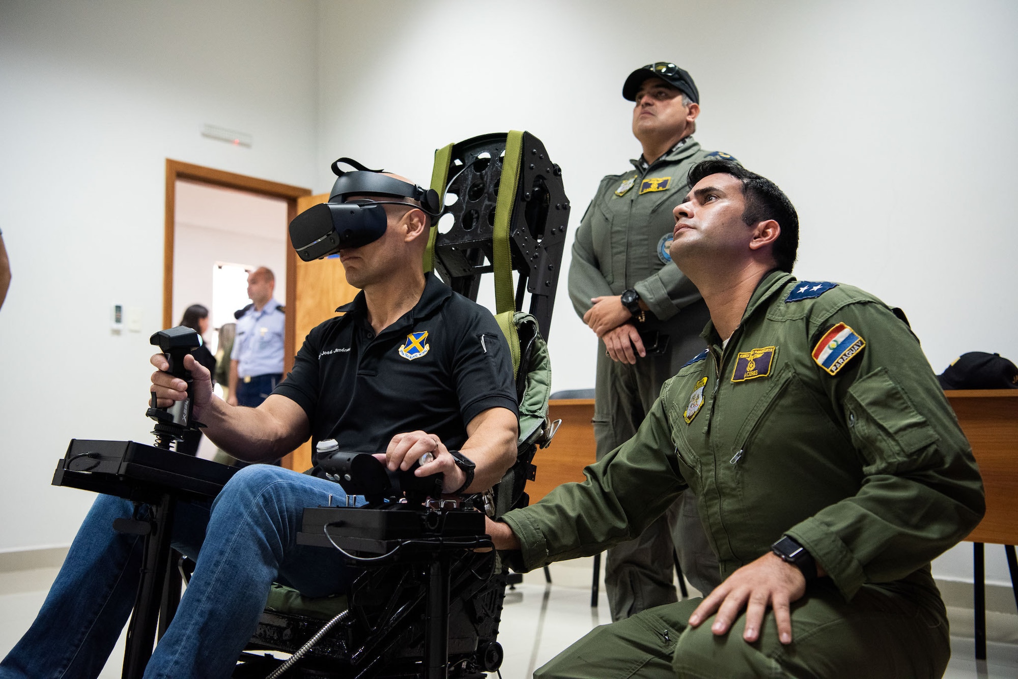 Two men sitting down looking up. Man on left is using virtual reality equipment while man on right is guiding him.