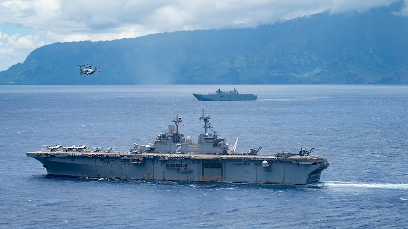 Two ships operate off the coast of Hawaii sail on the ocean as a helicopter hovers nearby. Mountains are in the background.