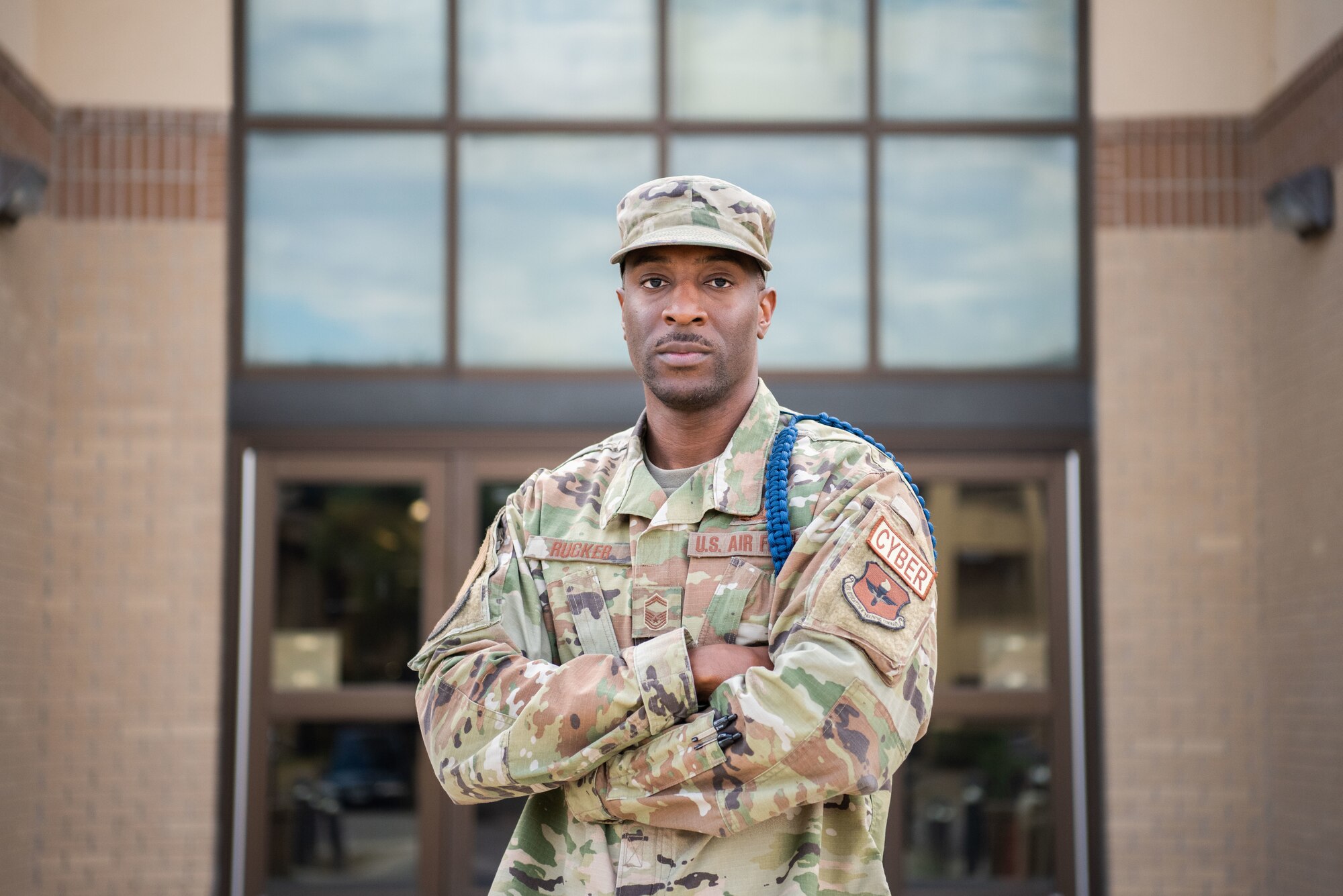 Man poses in front of a building, looking at the camera, with his arms folded across his body.