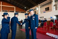 Four people frame the photo; the woman on the left is saluting the man positioned at the right side. The two people in the middle, both men, are shaking hands.