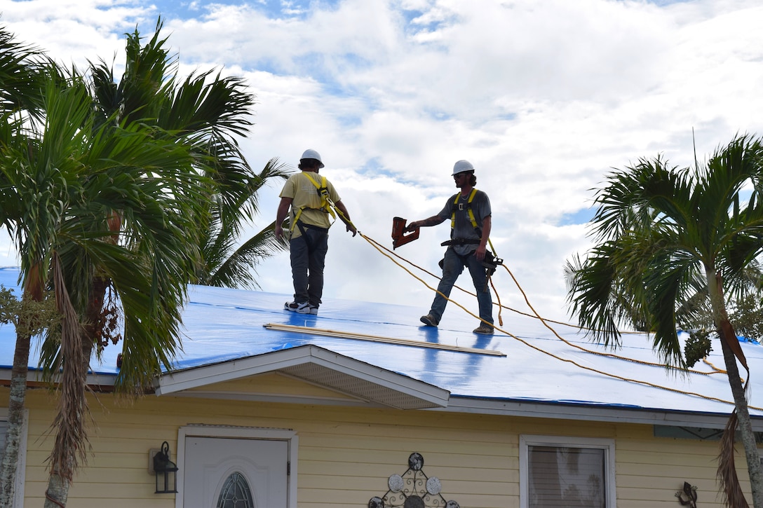 Two contractors wearing safety harnesses stand on a blue protective sheet on the roof of a home.