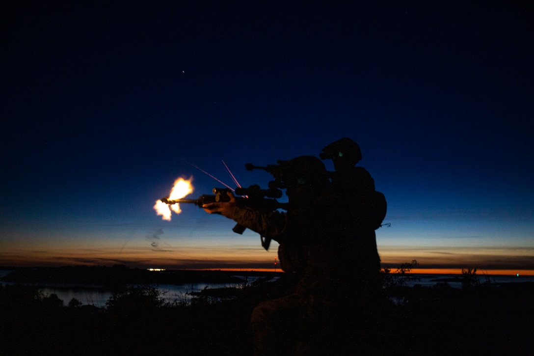 Marines fire weapons at night with a sliver of orange sky in the background.
