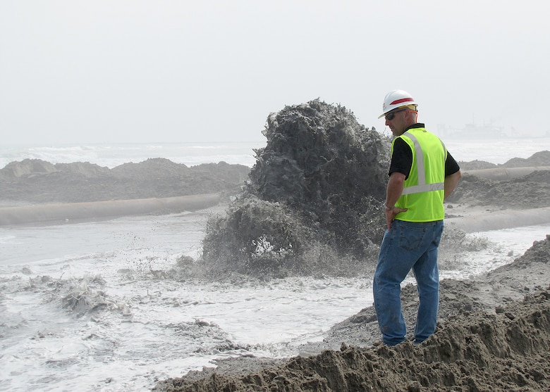 A man stands on a beach as waves crash upon the coastline. A grey plum from an explosion rises up from the water.