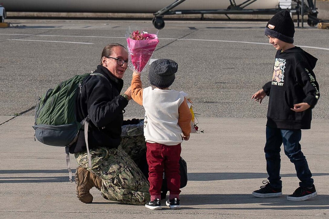 A sailor kneels on the ground as she is given flowers by a young child.