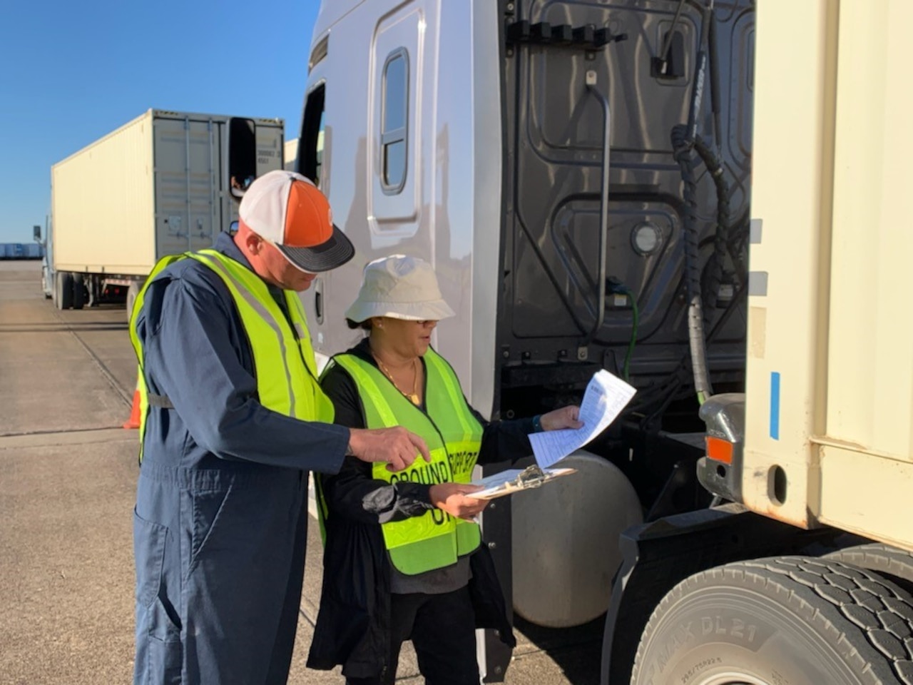 A man and woman wearing construction clothing stand next to a truck while looking at paperwork.