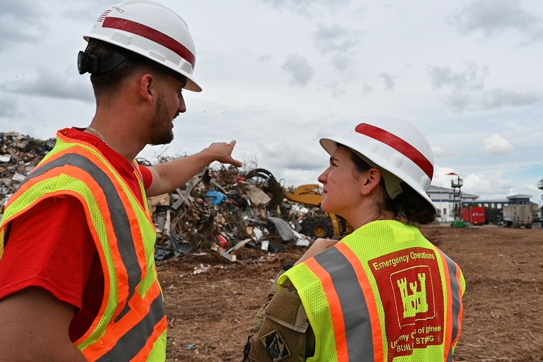 A man wearing a yellow reflective vest points at a large pile of debris while a woman who is also wearing a yellow reflective vest looks on.