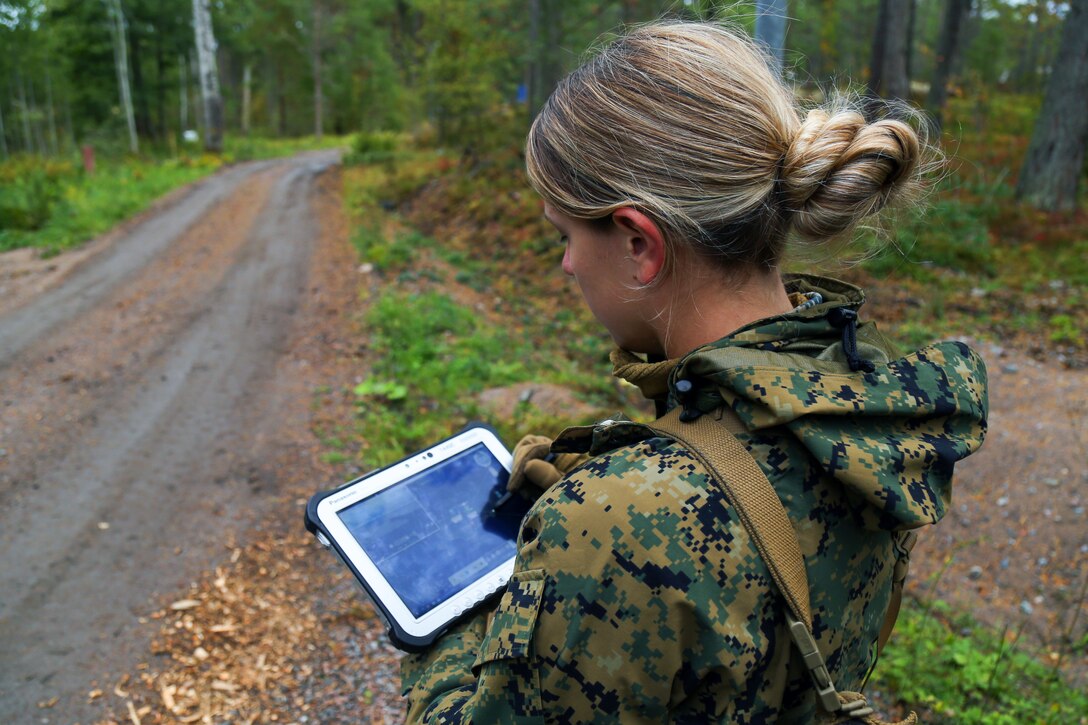 A Marine uses a stylus on a handheld device by a road in a forested area.