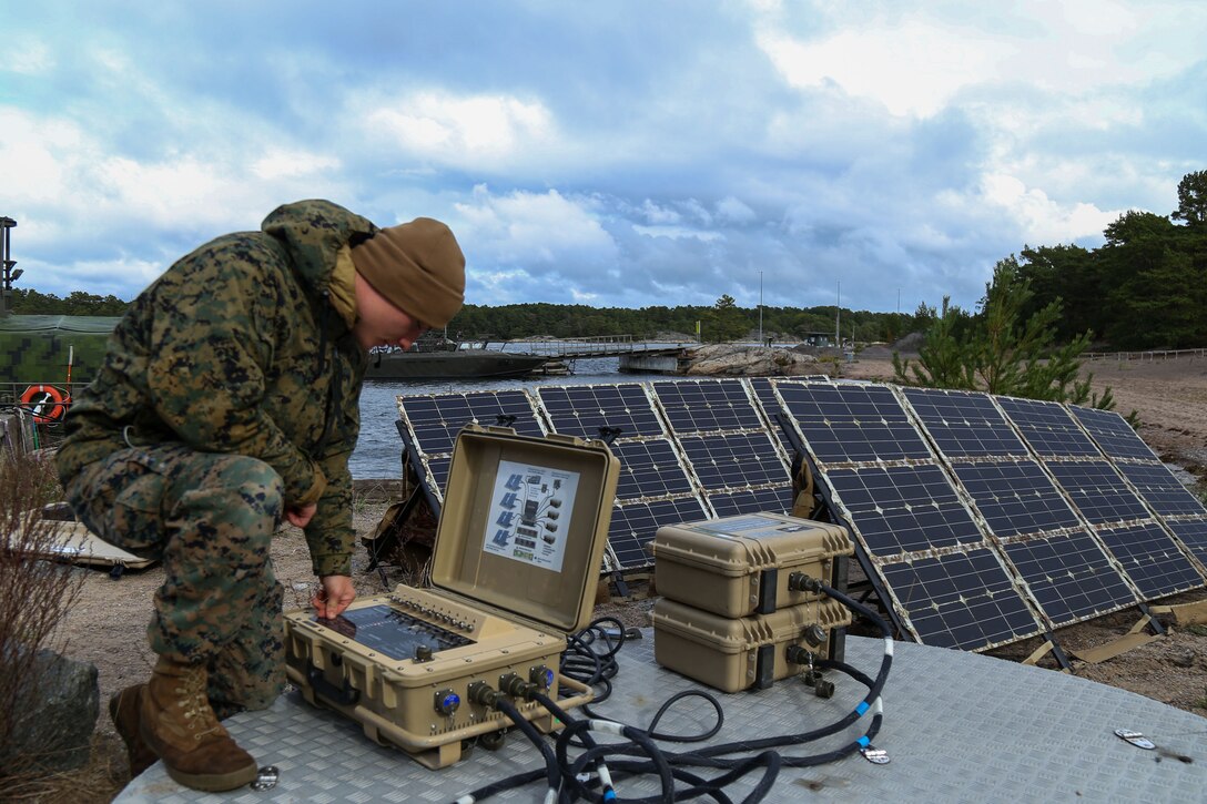 A Marine kneels over a device with cords coming out of it outside, with solar panels set up nearby.
