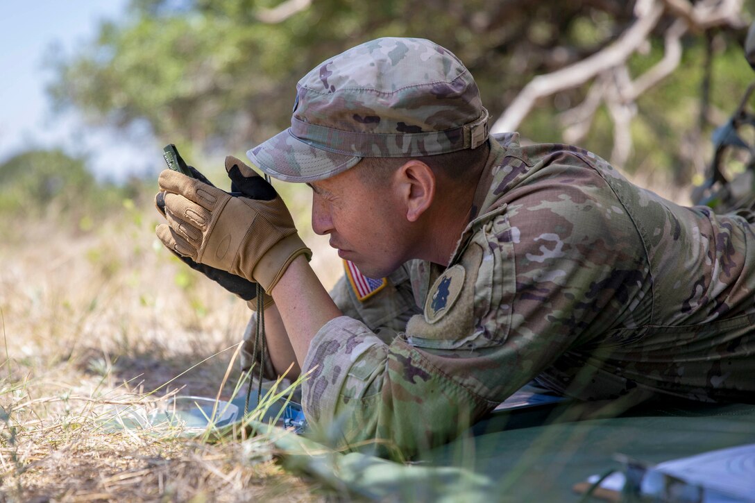 A soldier laying on the ground in a field uses a device to check the direction.