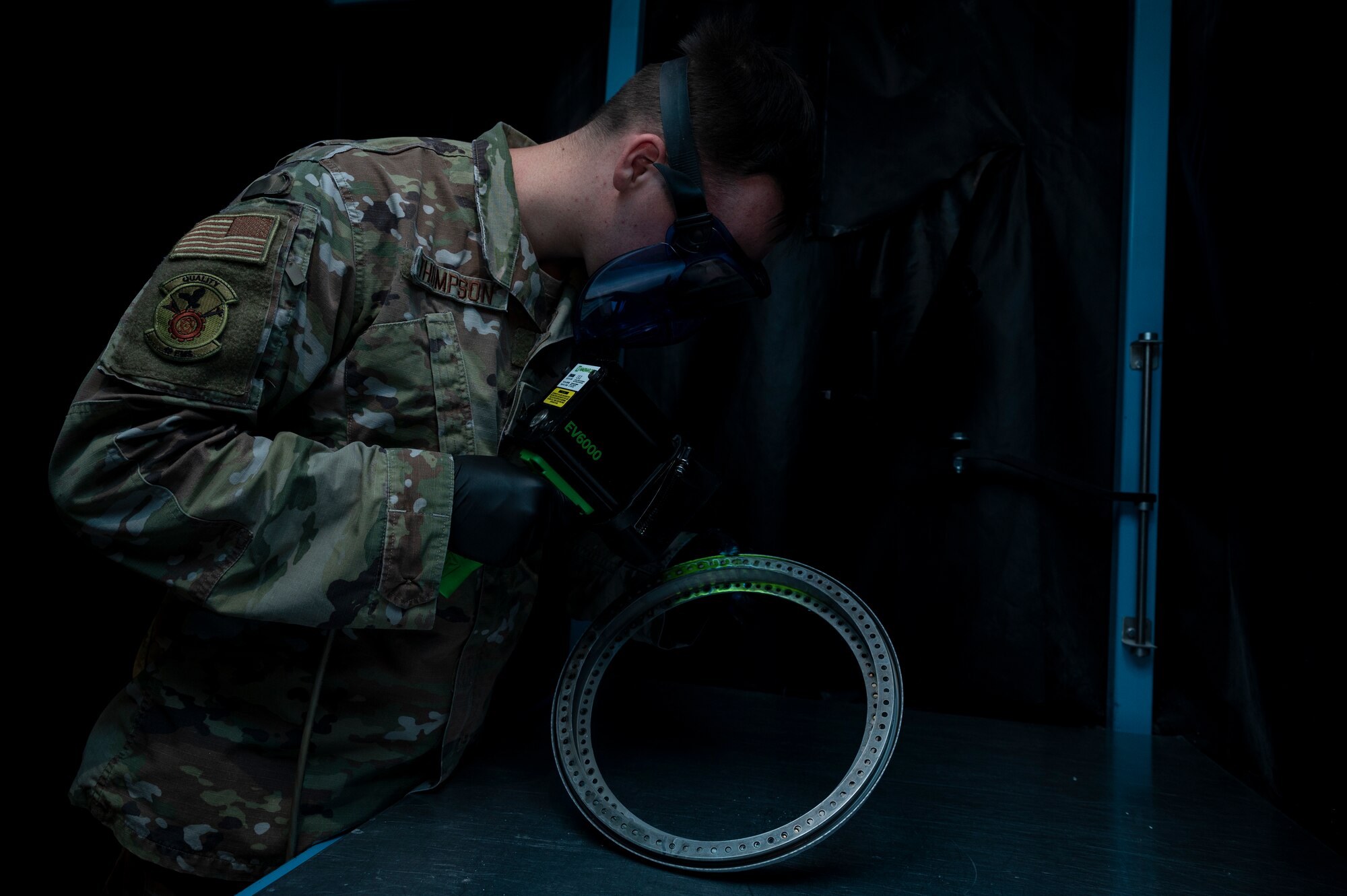 A man in green OCPs uses a black light to look over an aircraft part.