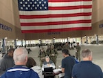 People lined up in front of a table with a person sitting down on it while looking at a laptop. A large group of uniformed military people are in the background.