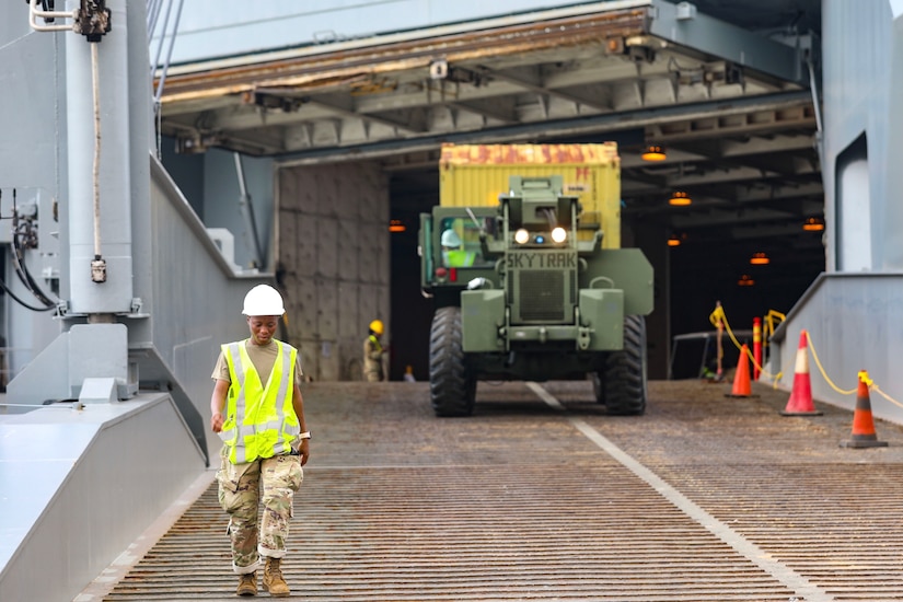 A service member walks in front of a military vehicle driving down a ramp off a ship.