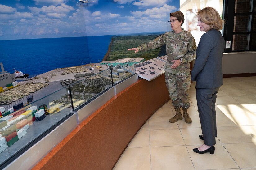 An airman gestures at a wall-sized illustration of a port scene while talking to a civilian.