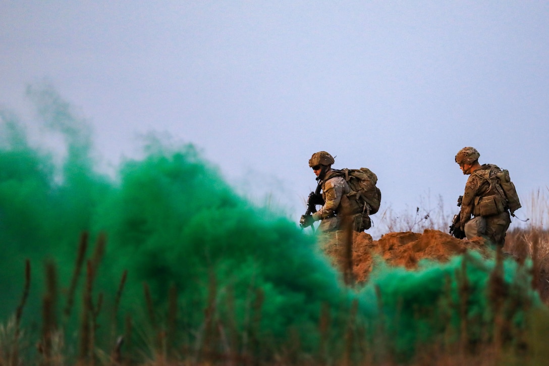 Two armed soldiers walk through a field behind a plume of green smoke.