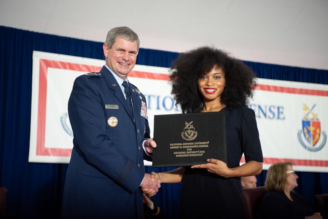 A man in a blue military dress uniform gives a diploma to a woman wearing a black dress