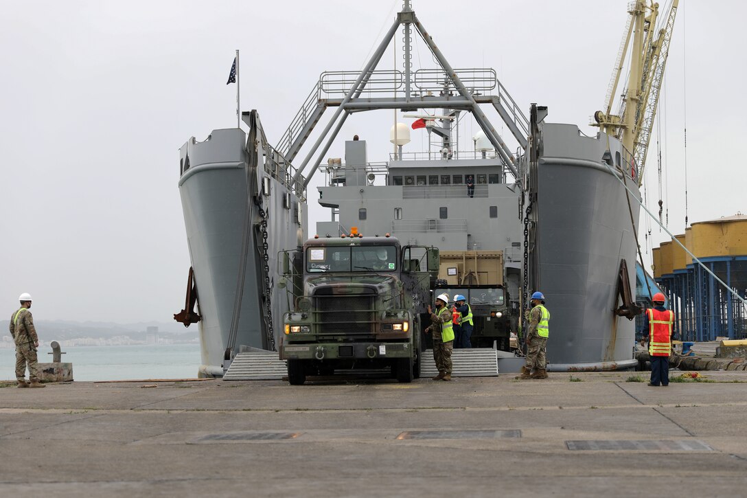 A person drives a military truck off of a ship’s ramp while several uniformed service members assist another person.