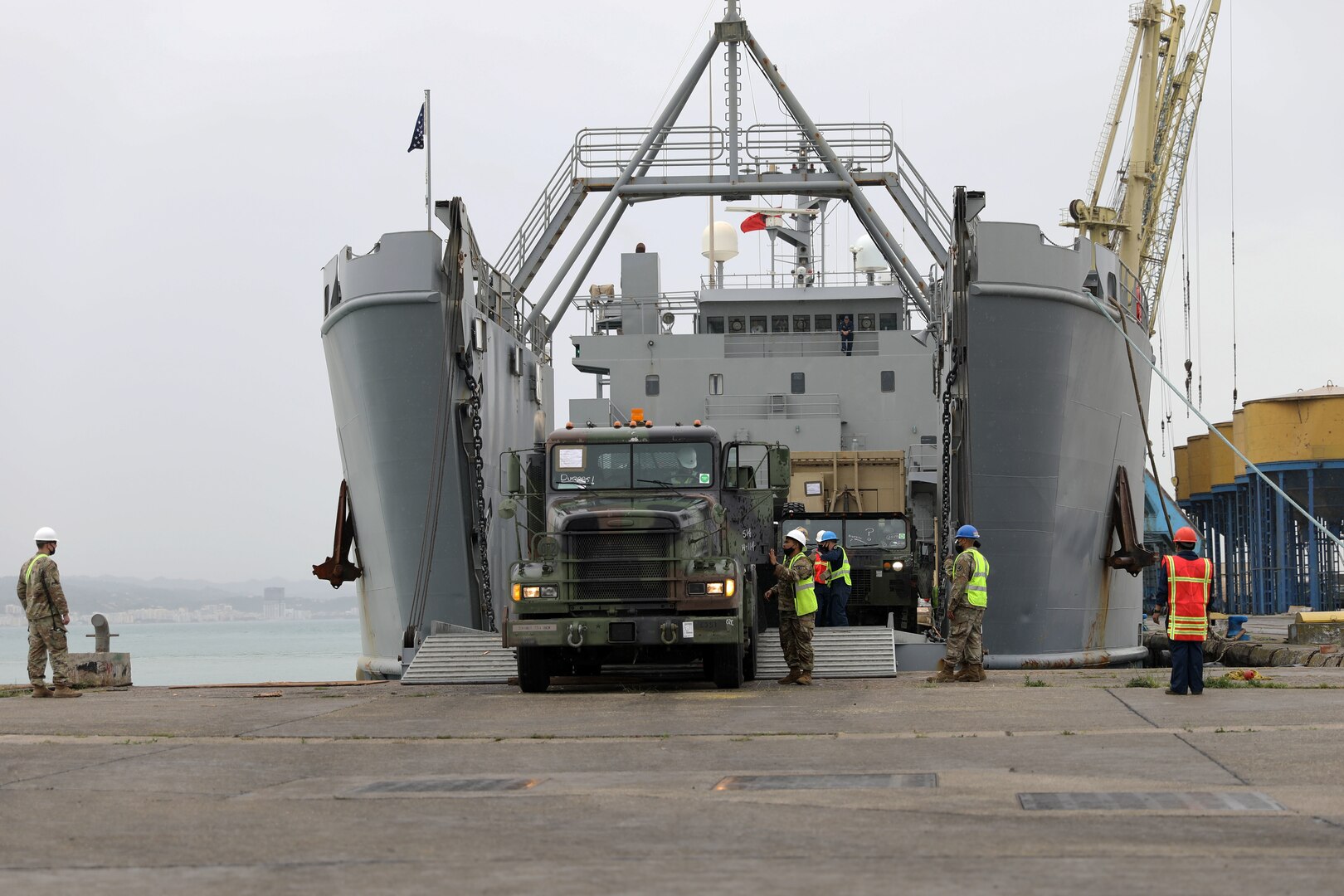 A person drives a military truck off of a ship’s ramp while several uniformed service members assist another person.