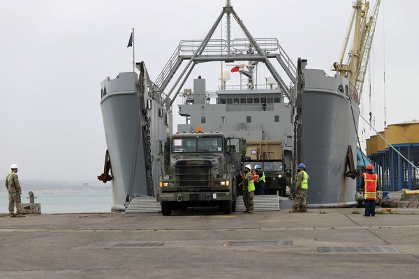 A person drives a military truck off of a ship’s ramp while several uniformed service members assist another person.