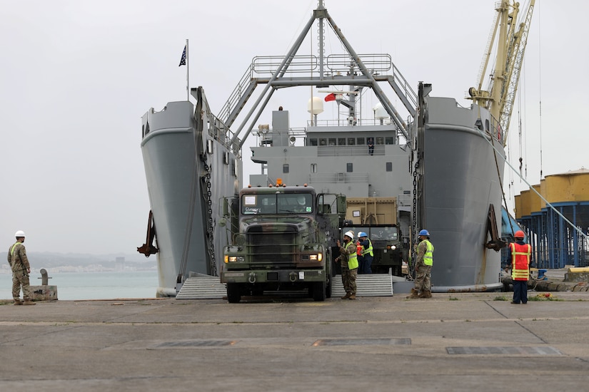 A person drives a military truck off of a ship’s ramp while several uniformed service members assist another person.
