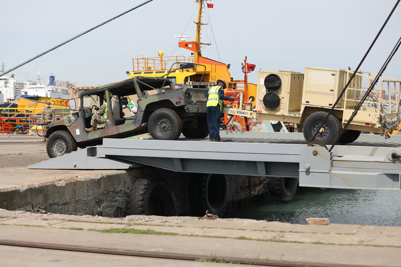 A uniformed service member drives a military tactical vehicle off a ship’s ramp.
