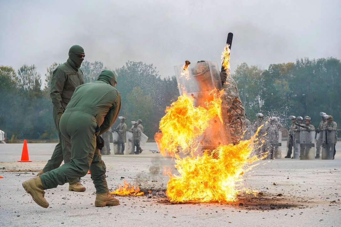 A soldier holding a clear shield walks through fire as fellow soldiers stand nearby.