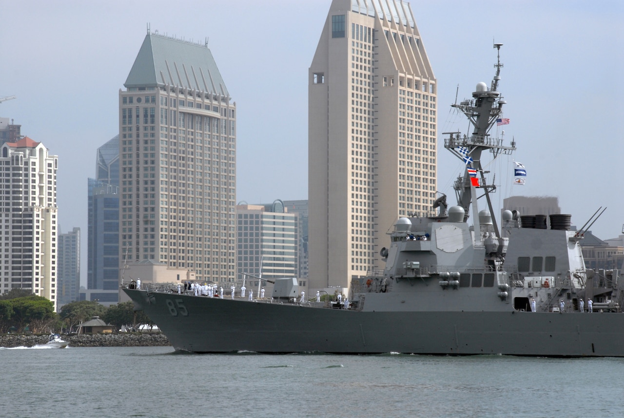 A destroyer ship sails in a harbor surrounded by high-rise buildings.
