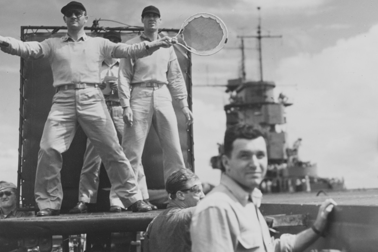 A man waves a paddle to help land a plane. Other men doing similar duties watch.