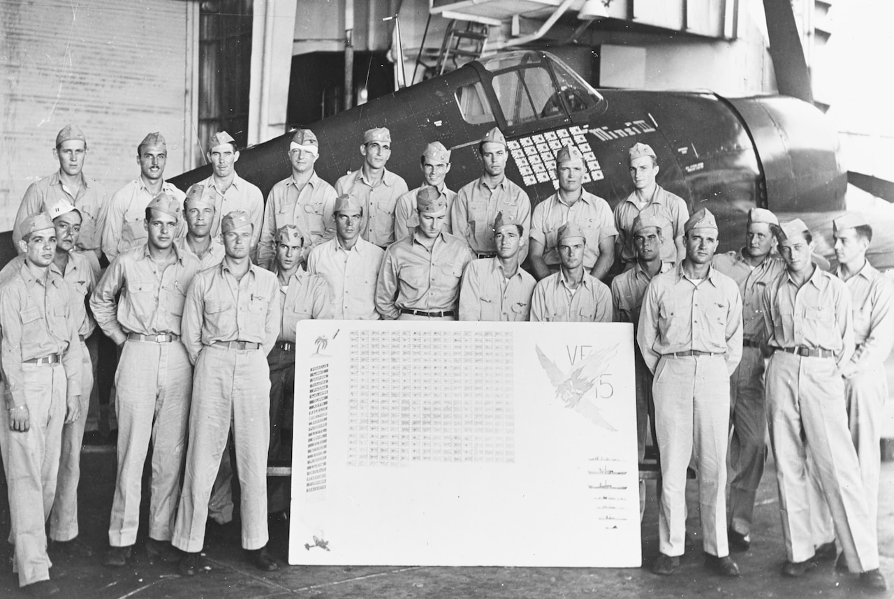 Two-dozen men stand around a scorecard and in front of a propeller airplane.