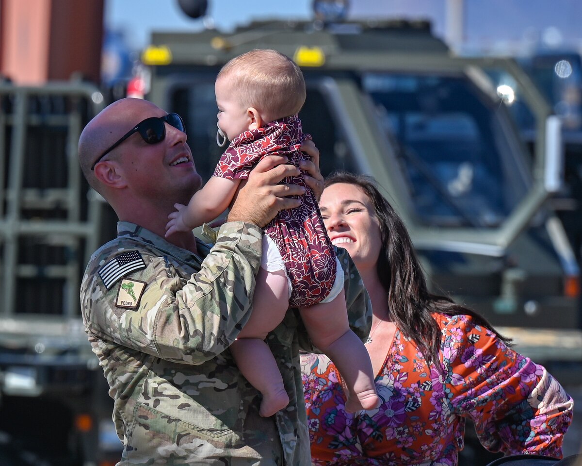 A photo of a man in uniform holding his young child while his wife smiles in the background.
