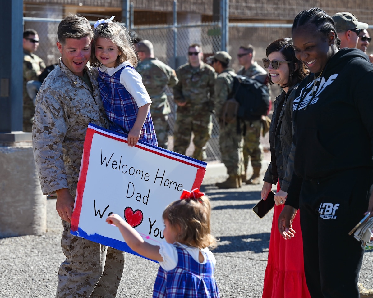 A photo of a man in uniform holding his daughter while his other daughter walks towards him.