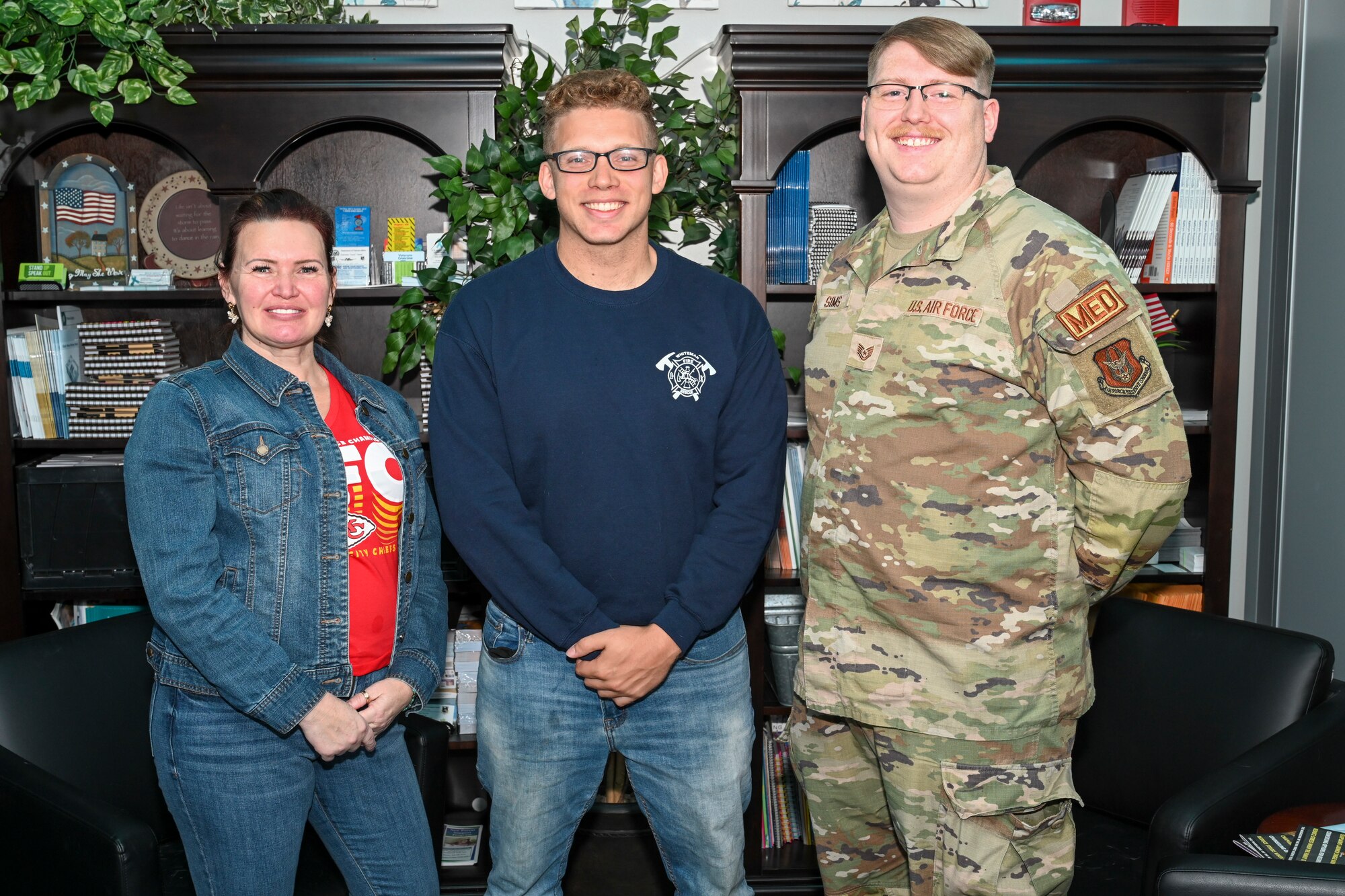 From left to right, a woman in a denim jacket and jeans, a man in jeans and a blue sweatshirt, and a man in camouflage fatigues smile at the camera in front of a pair of bookshelves.