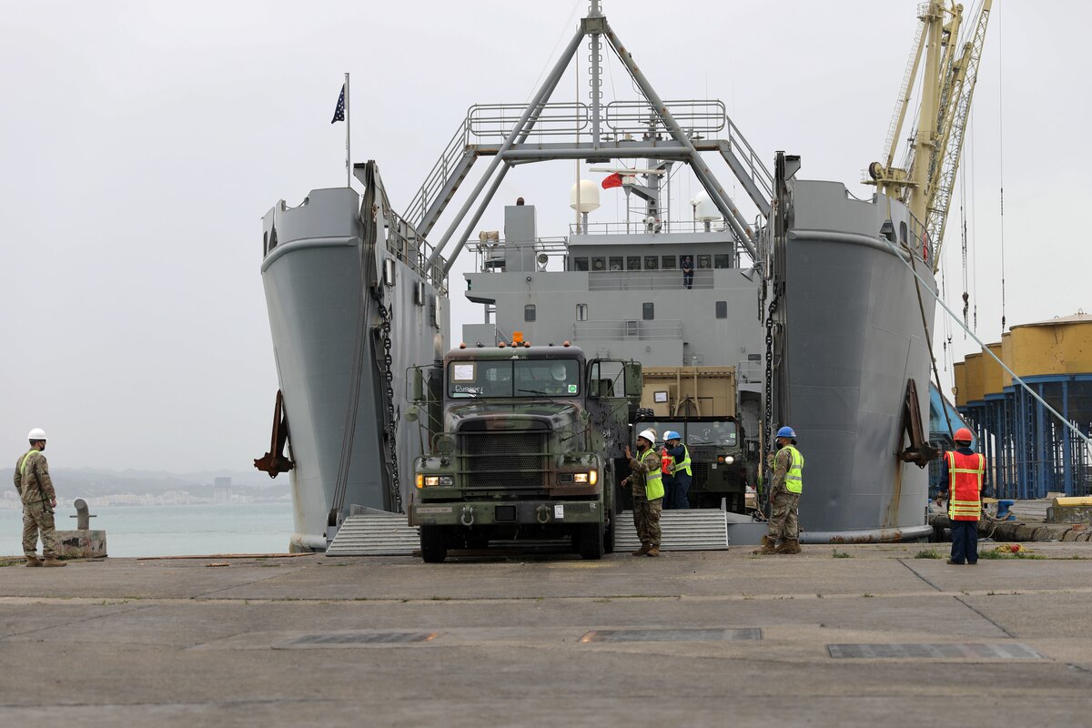 A person drives a military truck off of a ship’s ramp while several uniformed service members assist another person.