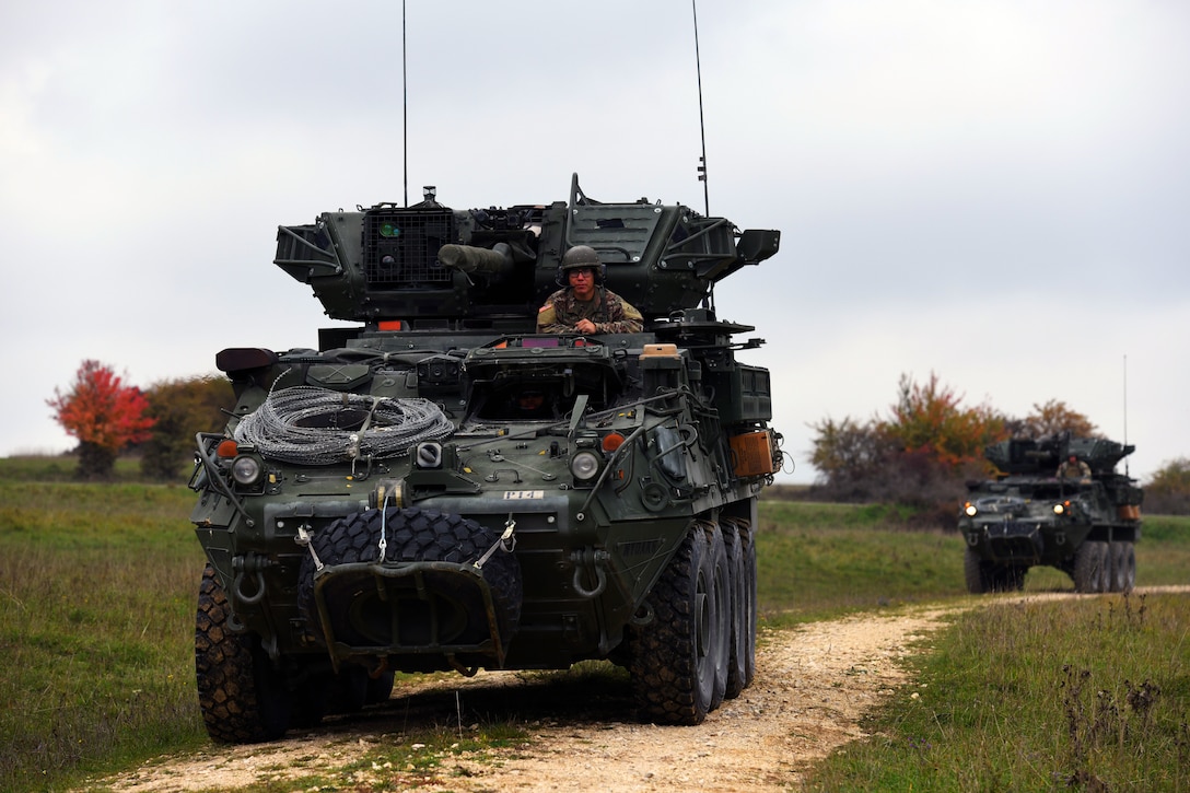Soldiers ride in two Stryker vehicles down a dirt path.
