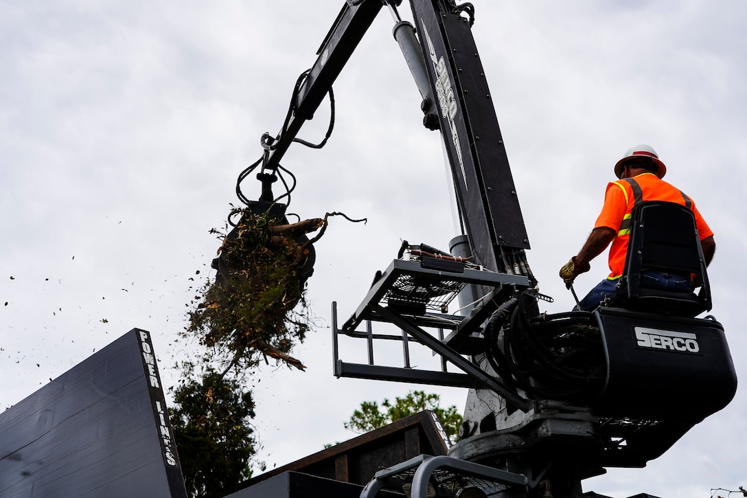 A man wearing an orange reflective vest operating a crane collects debris and places it in a compactor.