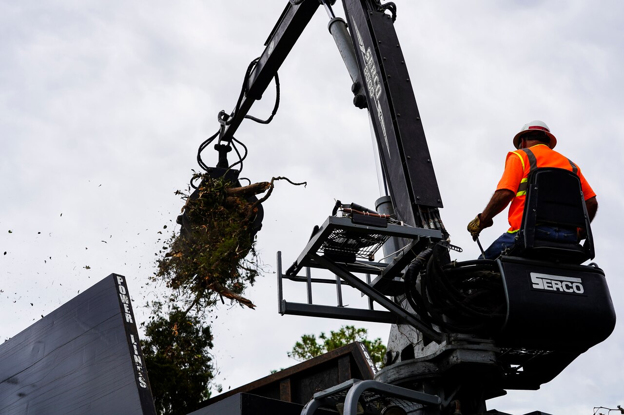 A man wearing an orange reflective vest operating a crane collects debris and places it in a compactor.