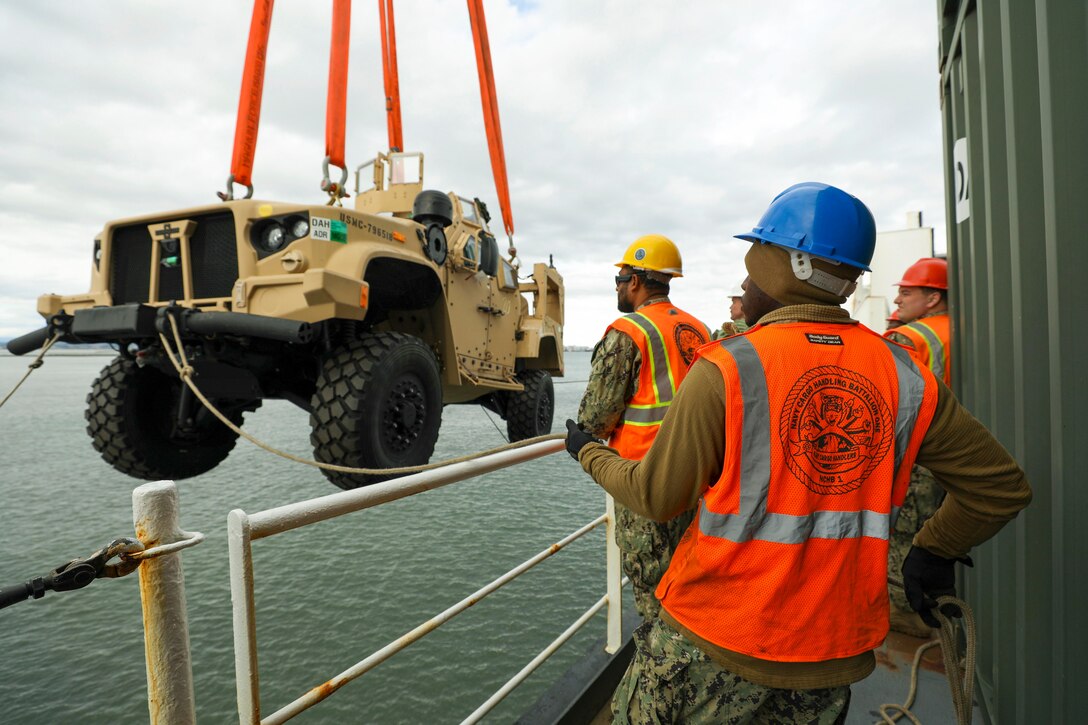 Sailors hold rope attached to a tactical vehicle being lifted onto a ship.