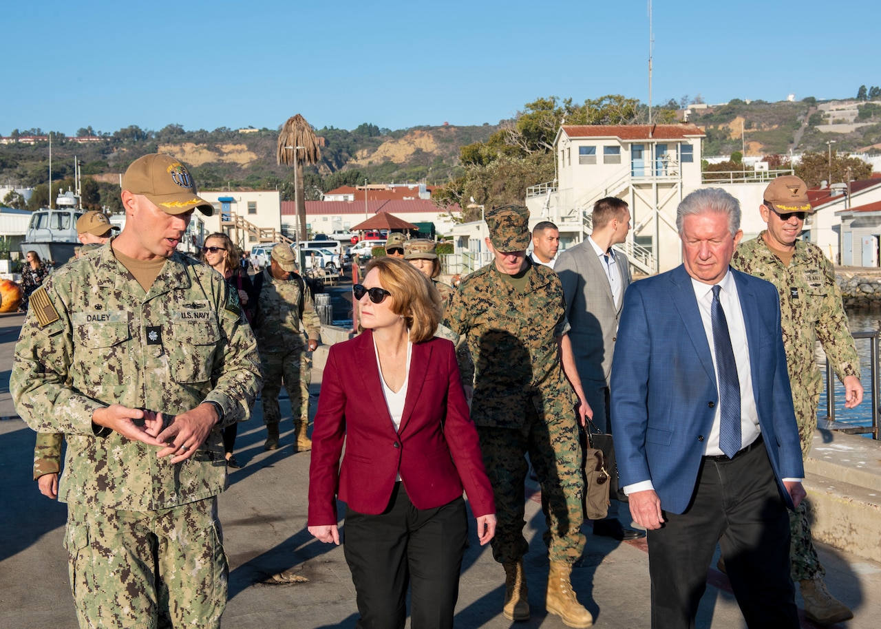 A man in a uniform and a woman in business attire walk past a boat and buildings with colleagues and service members.