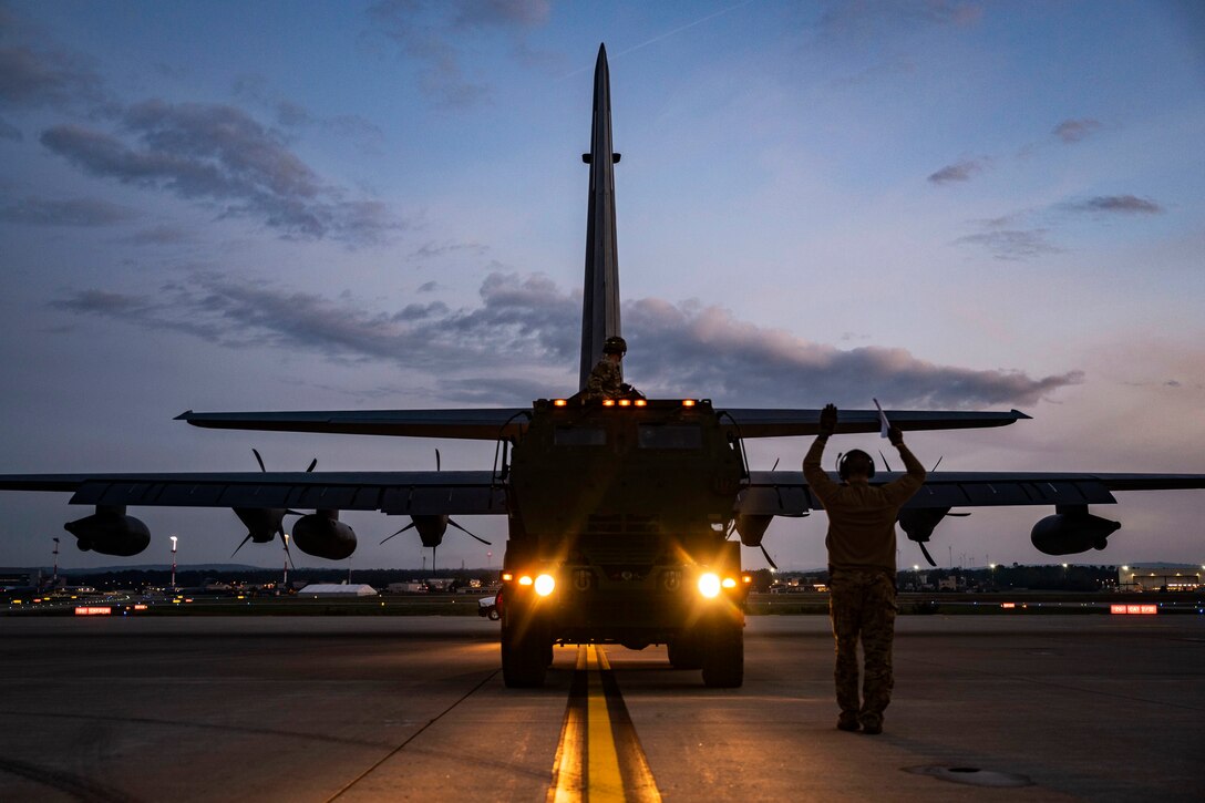 An airman holding wands signals toward a military vehicle and an aircraft.