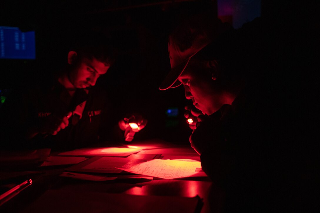 Two sailors use flashlights to look at documents in the dark.