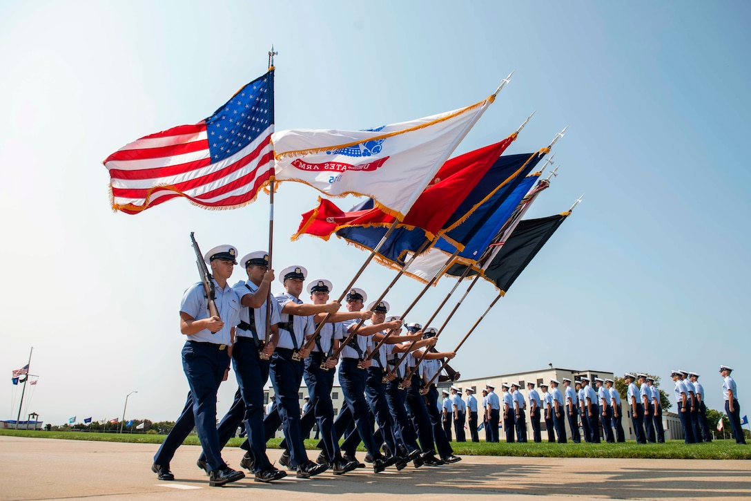 Coast Guard recruits walk in formation holding rifles and flags.